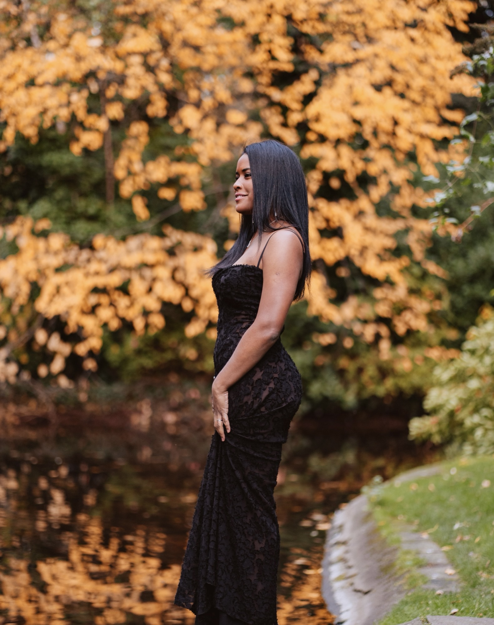 Full-body outdoor profile of Yazuri Guido in a black lace evening gown beside autumn foliage and water